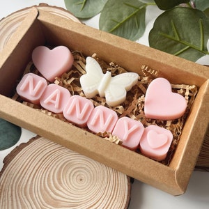 May include: A brown box filled with pink and white scented wax melts. The melts are shaped like hearts and letters, spelling out the word "NANNY". There is also a white butterfly shaped melt.