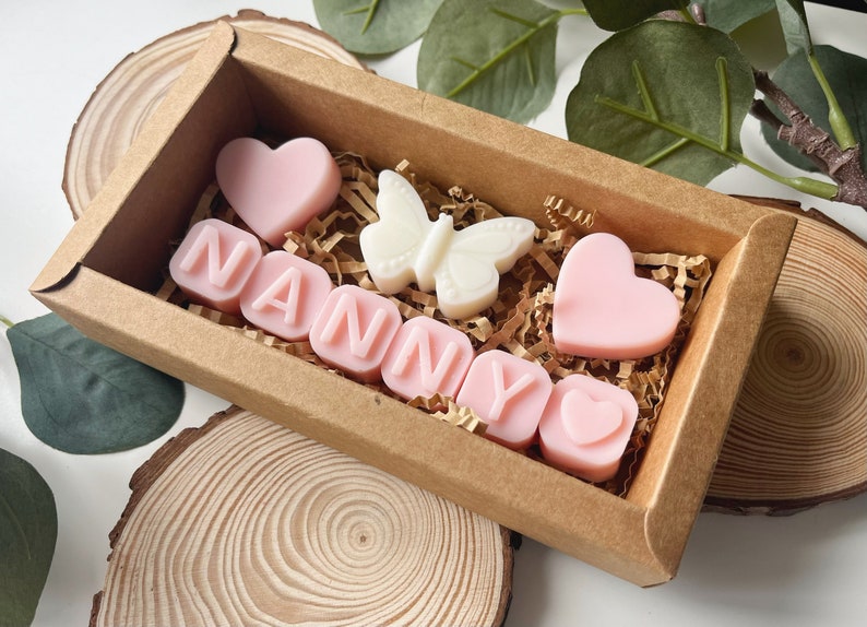 May include: A brown box filled with pink and white scented wax melts. The melts are shaped like hearts and letters, spelling out the word "NANNY". There is also a white butterfly shaped melt.