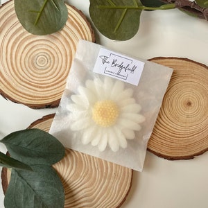 May include: A white daisy candle in a clear bag with the text "The Bridgefield Candle Company" on a white label. The candle is sitting on a wooden slice.