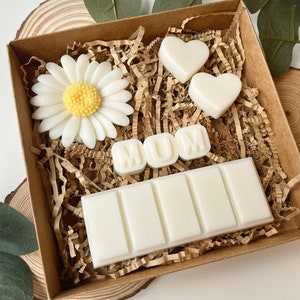 May include: A brown gift box filled with white wax melts. The melts are shaped like hearts, a daisy, and the letters "MUM". The box is filled with brown shredded paper.