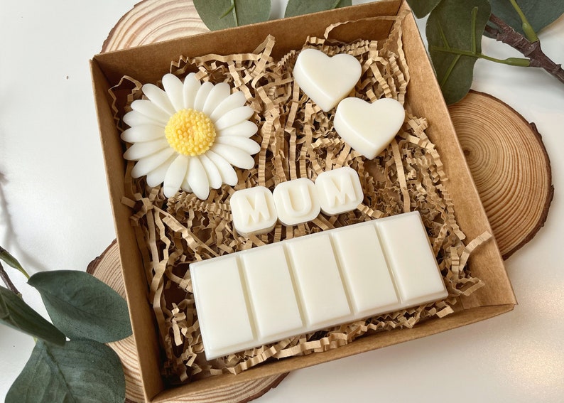May include: A brown gift box filled with white wax melts. The melts are shaped like hearts, a daisy, and the letters "MUM". The box is filled with brown shredded paper.