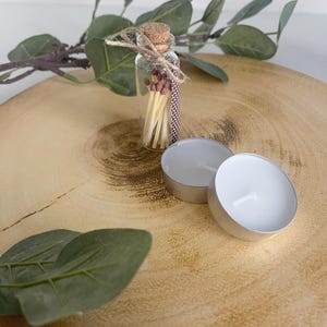 May include: A small clear glass bottle with a cork stopper containing wooden matches with brown tips, tied with a piece of twine. Next to the bottle are two round white tea light candles in silver metal cups, accompanied by green leaves.