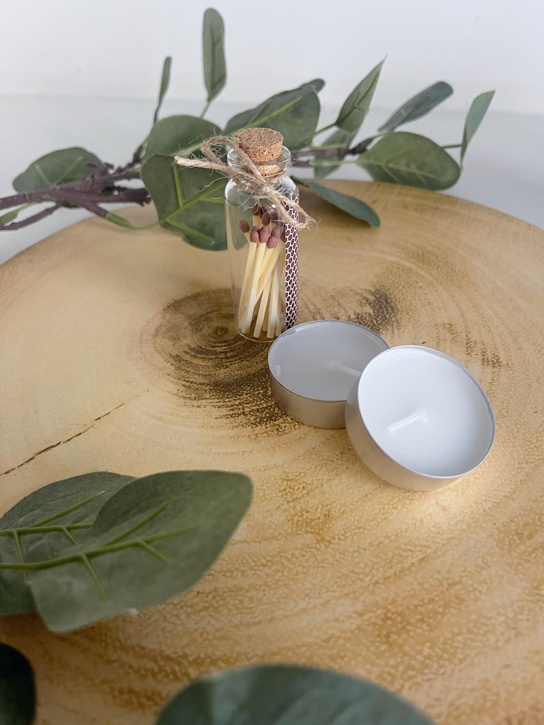 May include: A small clear glass bottle with a cork stopper containing wooden matches with brown tips, tied with a piece of twine. Next to the bottle are two round white tea light candles in silver metal cups, accompanied by green leaves.