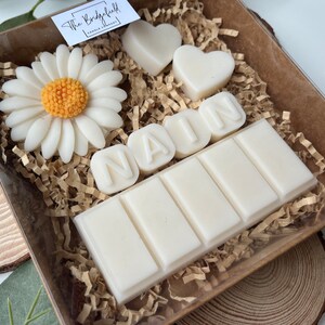 May include: A white gift box filled with white wax melts, including a daisy, two hearts, and the name "NAIM" spelled out in individual letters. The box is lined with brown shredded paper.