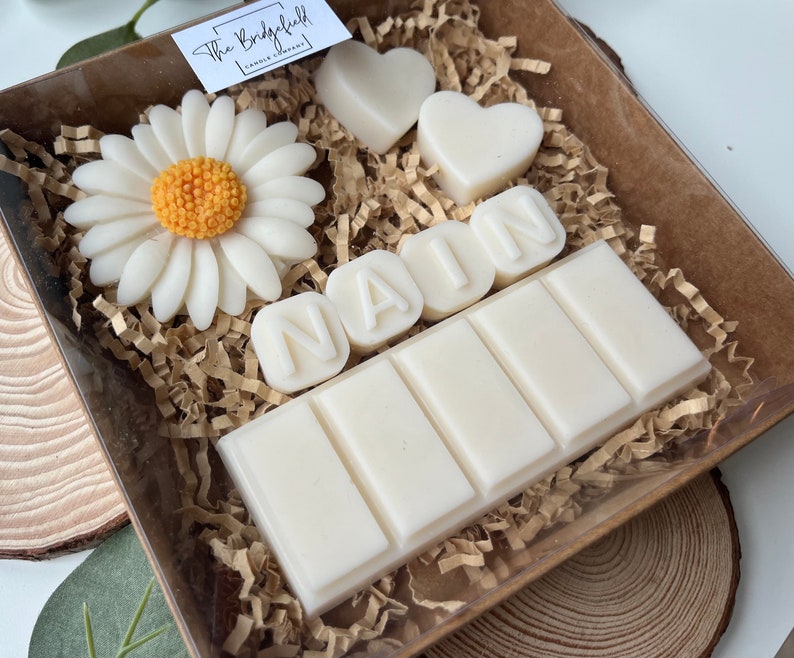 May include: A white gift box filled with white wax melts, including a daisy, two hearts, and the name "NAIM" spelled out in individual letters. The box is lined with brown shredded paper.