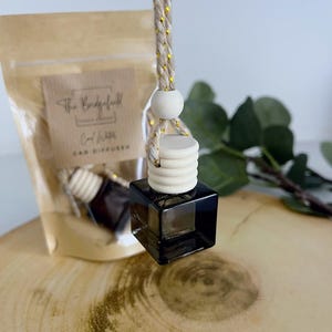 May include: A black square car air freshener with a wooden top and a beige and gold rope. A clear plastic bag with the text "The Bridgefield" and "Cool Waters" is in the background. The car diffuser is on a wooden surface.