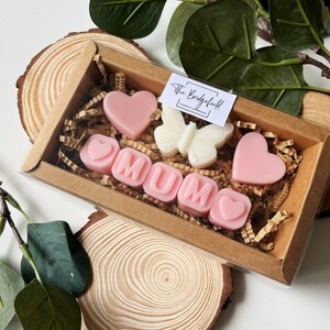 May include: A gift box filled with pink and white wax melts, spelling out "MUM" with hearts and a butterfly. The box is made of brown cardboard and has a clear plastic window.