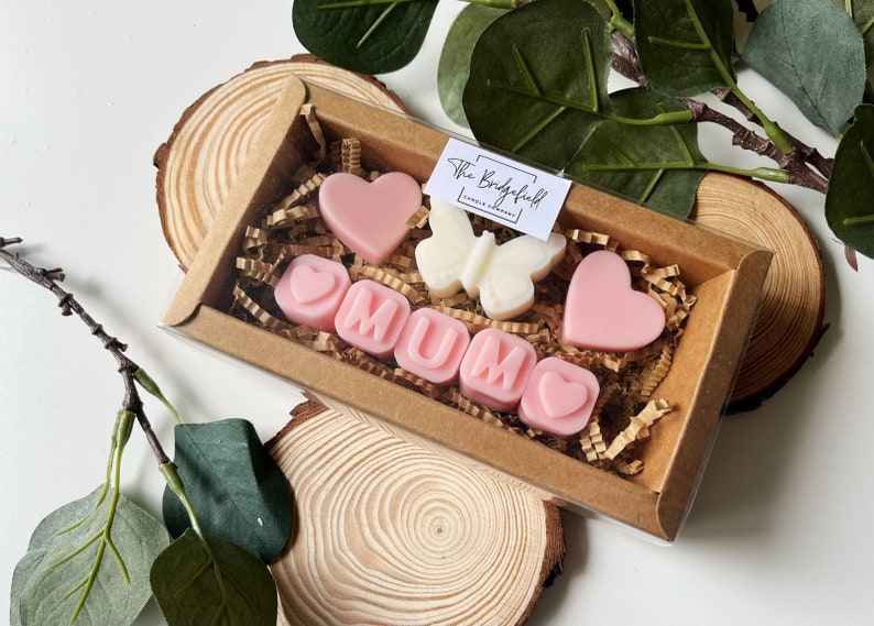 May include: A gift box filled with pink and white wax melts, spelling out "MUM" with hearts and a butterfly. The box is made of brown cardboard and has a clear plastic window.
