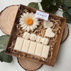 May include: A gift box filled with white wax melts, including a daisy, two hearts, and the name "NAIN" spelled out. The box is lined with brown shredded paper and has a label that reads "The Bridgefield Candle Company".