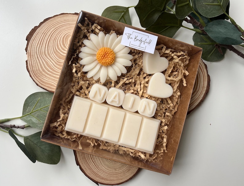 May include: A gift box filled with white wax melts, including a daisy, two hearts, and the name "NAIN" spelled out. The box is lined with brown shredded paper and has a label that reads "The Bridgefield Candle Company".