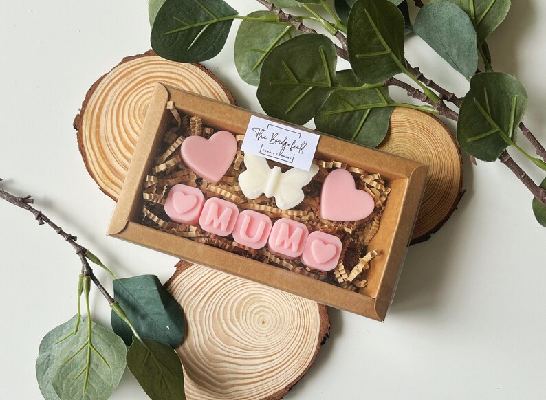 May include: A gift box filled with pink and white wax melts shaped like hearts and the word "MUM". The box is on a wooden surface with green leaves.