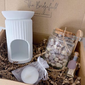 May include: A white ceramic wax warmer, a glass jar filled with small wax melts, a small glass bottle of matches, and a tea light candle in a white organza bag. The items are packaged in a cardboard box with The Bridgefield Candle Company logo.