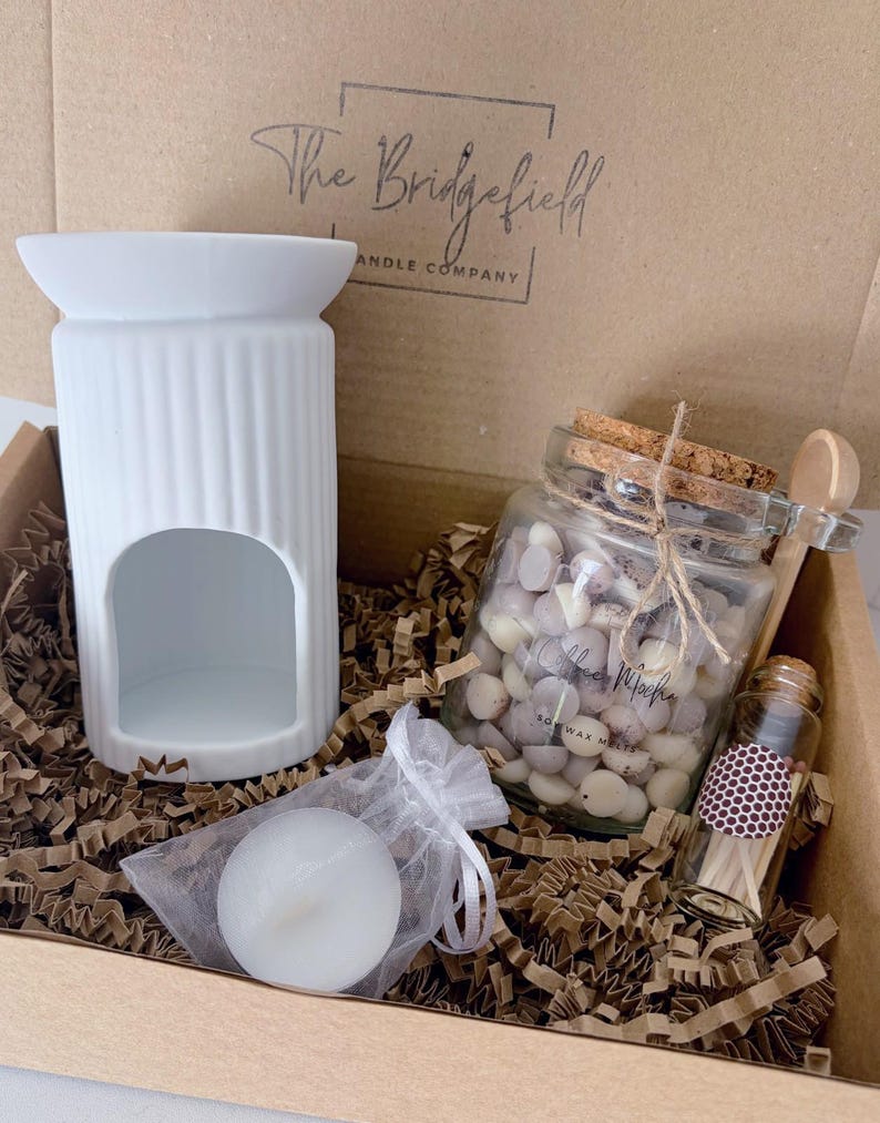 May include: A white ceramic wax warmer, a glass jar filled with small wax melts, a small glass bottle of matches, and a tea light candle in a white organza bag. The items are packaged in a cardboard box with The Bridgefield Candle Company logo.