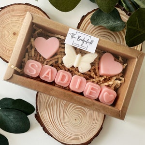 May include: A brown box filled with pink and white wax melts. The melts are shaped like hearts, a butterfly, and the letters S-A-D-I-E. The box has a label that says "The Bridgefield Candle Company."