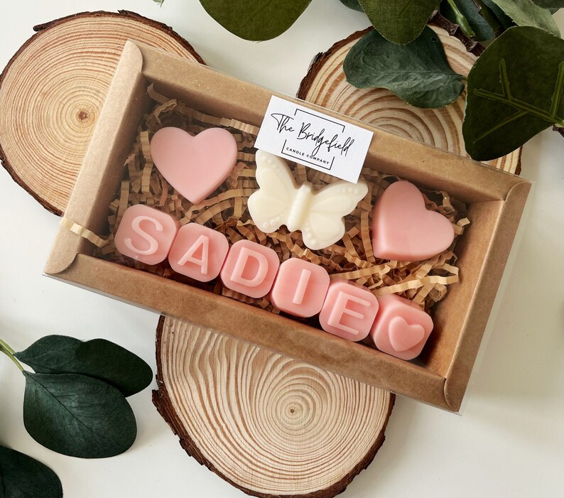 May include: A brown box filled with pink and white wax melts. The melts are shaped like hearts, a butterfly, and the letters S-A-D-I-E. The box has a label that says "The Bridgefield Candle Company."