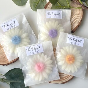 May include: Four white wax daisy candles with different colored centers. The candles are packaged in individual white tissue paper bags with a label that reads "The Bridgefield Candle Company".