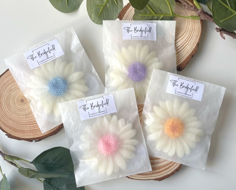 May include: Four white wax daisy candles with different colored centers. The candles are packaged in individual white tissue paper bags with a label that reads "The Bridgefield Candle Company".