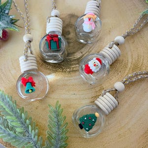 May include: Five clear glass Christmas-themed car air fresheners with wooden tops and decorative charms. Charms include a gift, snowman, Santa, Christmas tree, and a bell. Each freshener has a braided cord for hanging.