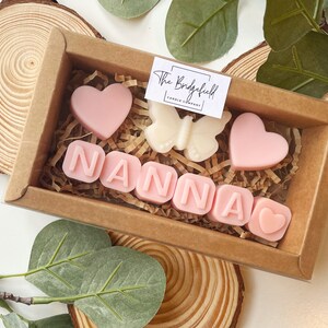 May include: A small gift box filled with pink and white scented wax melts. The melts are shaped like hearts and letters, spelling out the word "NANNA". The box is made of brown cardboard and has a clear plastic window.