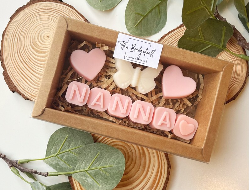 May include: A small gift box filled with pink and white scented wax melts. The melts are shaped like hearts and letters, spelling out the word "NANNA". The box is made of brown cardboard and has a clear plastic window.