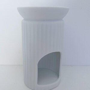 May include: A matte white ceramic wax warmer featuring a cylindrical body with vertical ribbed detailing. The top section has a wide, shallow basin for wax melts, while the lower portion includes a large arched opening for a tea light candle.