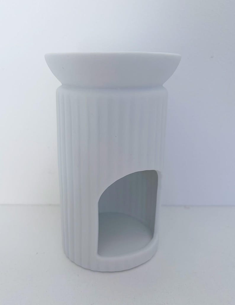 May include: A matte white ceramic wax warmer featuring a cylindrical body with vertical ribbed detailing. The top section has a wide, shallow basin for wax melts, while the lower portion includes a large arched opening for a tea light candle.