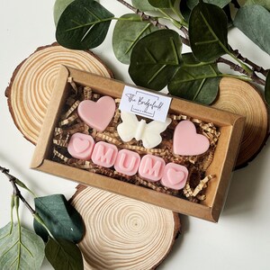 May include: A brown box filled with pink and white wax melts shaped like hearts and letters spelling "MUM". The box is sitting on a wooden surface. The wax melts are from The Bridgefield Candle Company.