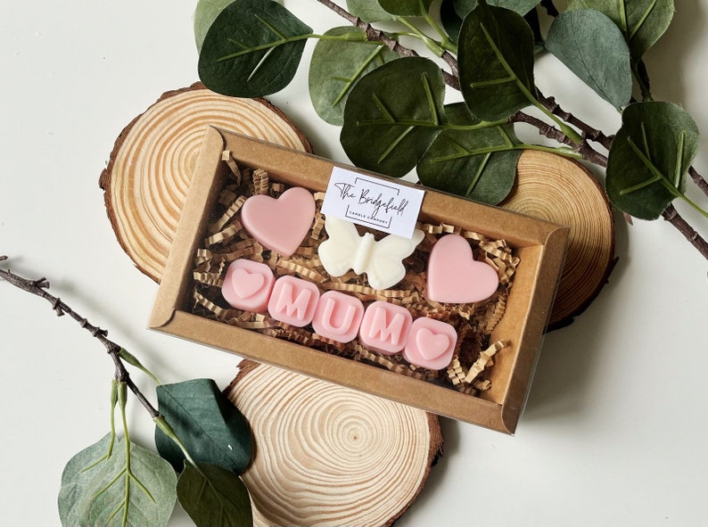 May include: A brown box filled with pink and white wax melts shaped like hearts and letters spelling "MUM". The box is sitting on a wooden surface. The wax melts are from The Bridgefield Candle Company.