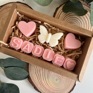 May include: A small brown box filled with pink and white scented wax melts. The melts are shaped like hearts and letters, spelling out the name "SADIE". There is also a white butterfly shaped melt.