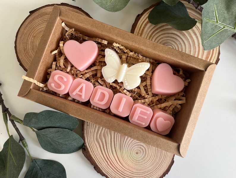 May include: A small brown box filled with pink and white scented wax melts. The melts are shaped like hearts and letters, spelling out the name "SADIE". There is also a white butterfly shaped melt.