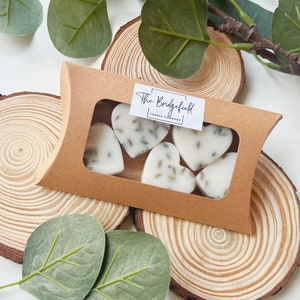 May include: A brown cardboard box with a window showing six heart-shaped wax melts. The white melts have lavender buds. The box has a label reading "The Bridgefield Candle Company". The box is on a wooden slice, with eucalyptus leaves.
