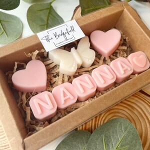 May include: A gift box filled with pink and white scented wax melts. The melts are shaped like hearts and letters, spelling out the word "NANNA". The box is decorated with a small white butterfly wax melt.