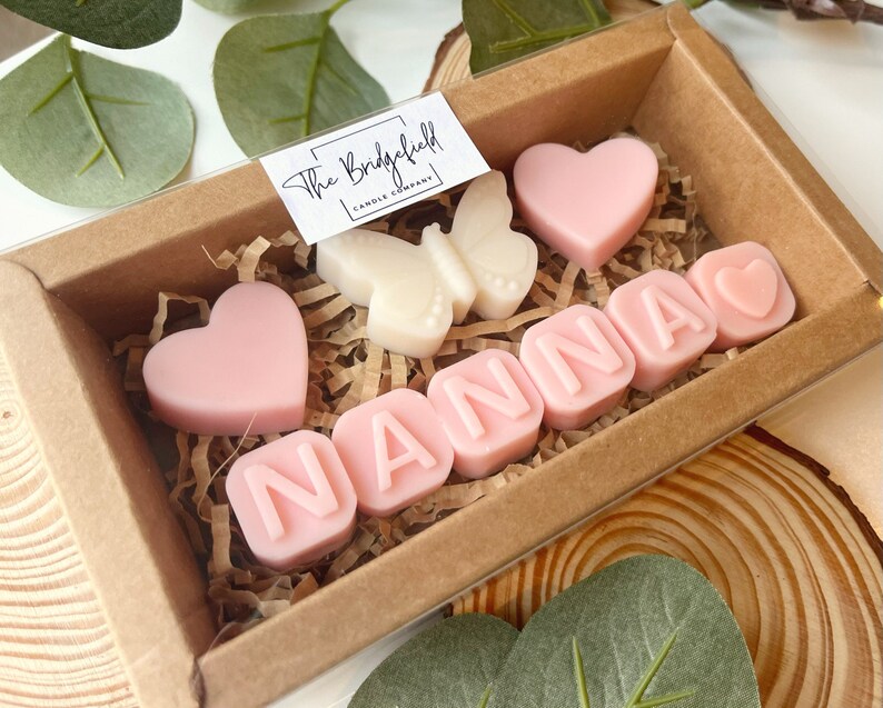 May include: A gift box filled with pink and white scented wax melts. The melts are shaped like hearts and letters, spelling out the word "NANNA". The box is decorated with a small white butterfly wax melt.