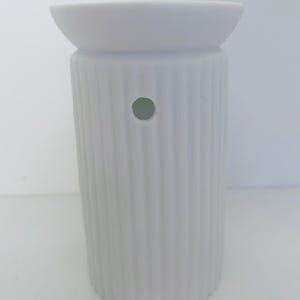 May include: A matte white ceramic wax warmer featuring a cylindrical body with vertical ribbed detailing. The top section has a wide, shallow basin for wax melts, and the side of the main body includes a single circular ventilation hole.