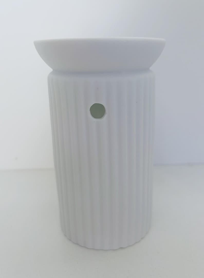 May include: A matte white ceramic wax warmer featuring a cylindrical body with vertical ribbed detailing. The top section has a wide, shallow basin for wax melts, and the side of the main body includes a single circular ventilation hole.