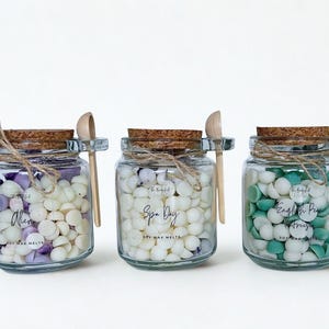 May include: Three clear glass jars filled with small, hemispherical soy wax melts. Each jar has a cork lid, a small wooden spoon attached with twine, and a label reading Alien, Spa Day, or English Pear & Freesia. The wax melts are white, purple, and green.