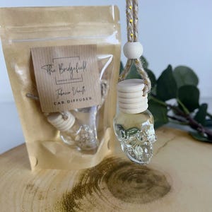 May include: A clear glass skull-shaped car air freshener with a wooden cap hangs from a gold and beige rope. A small pouch with a label reading "The Bridgefield Candle Company" and "Tobacco Vanilla Car Diffuser" is in the background.