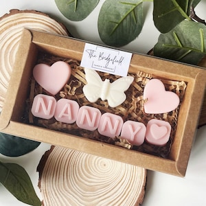 May include: A brown box filled with pink and white scented wax melts. The melts are shaped like hearts and letters, spelling out the word "NANNY". There is also a white butterfly shaped melt. The box is sitting on a wooden surface.