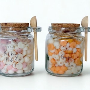 May include: Four clear glass jars filled with small, round wax melts in shades of pink, white, orange, and yellow. Each jar has a cork lid, a small wooden scoop tied with twine, and a label reading Soy Wax Melts with specific scent names.