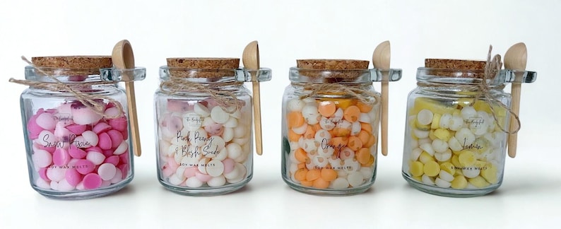 May include: Four clear glass jars filled with small, round wax melts in shades of pink, white, orange, and yellow. Each jar has a cork lid, a small wooden scoop tied with twine, and a label reading Soy Wax Melts with specific scent names.