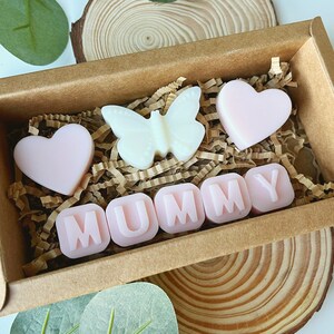 May include: A brown box filled with pink and white wax melts. The melts are shaped like hearts, a butterfly, and the word "MUMMY".