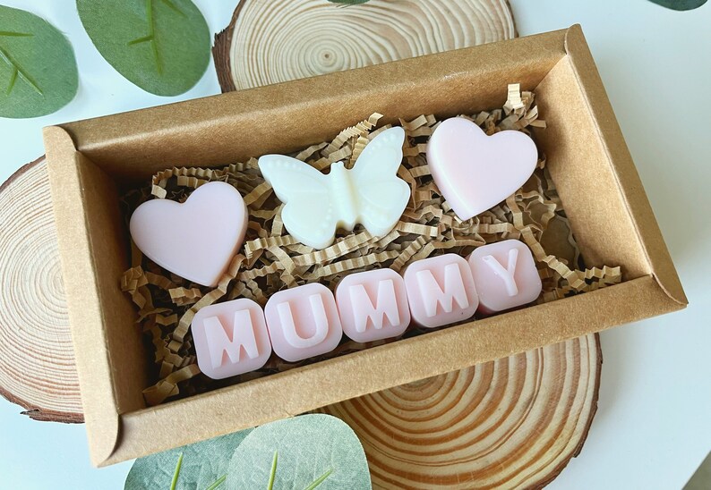 May include: A brown box filled with pink and white wax melts. The melts are shaped like hearts, a butterfly, and the word "MUMMY".