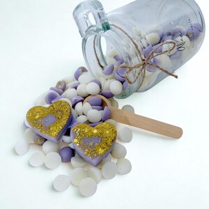 May include: A clear glass jar tipped on its side, spilling small white and lavender wax melts. Two heart-shaped wax melts with gold glitter accents rest in the pile. A wooden scoop lies among the wax pieces. The jar features a twine bow and text.