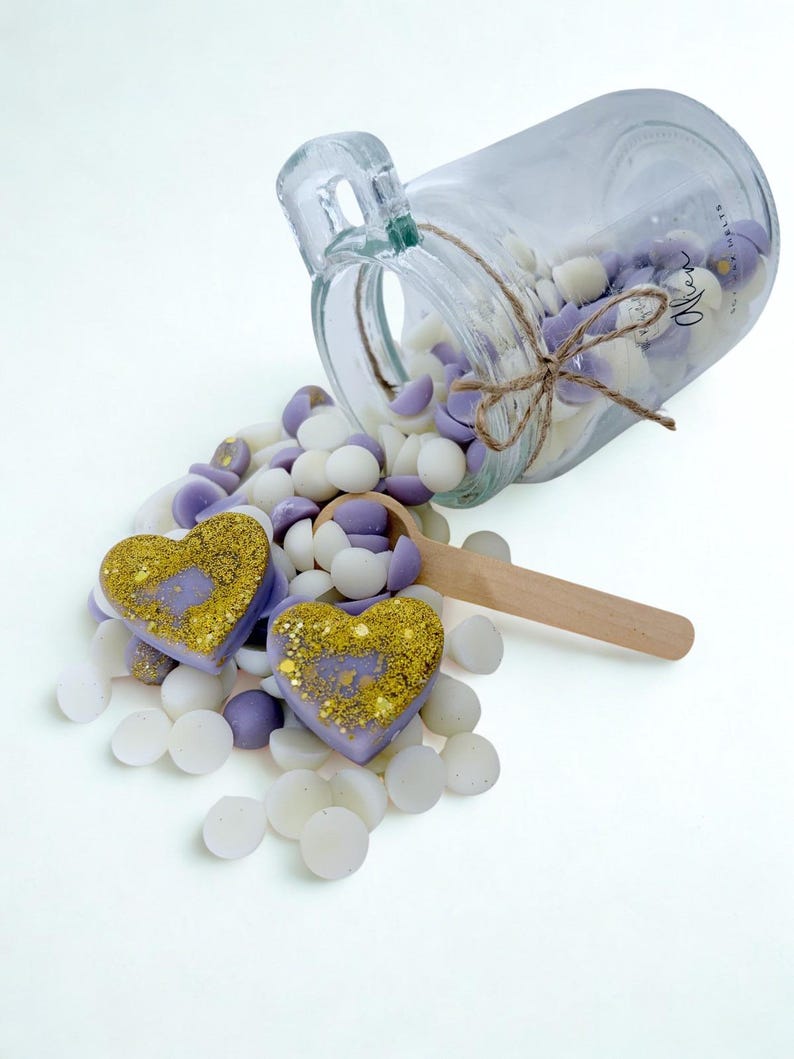 May include: A clear glass jar tipped on its side, spilling small white and lavender wax melts. Two heart-shaped wax melts with gold glitter accents rest in the pile. A wooden scoop lies among the wax pieces. The jar features a twine bow and text.