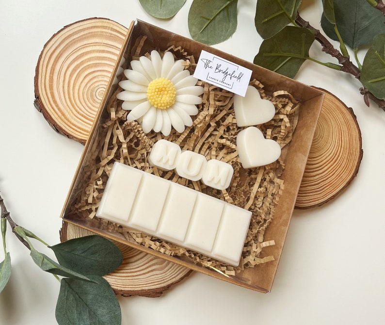 May include: A gift box filled with white wax melts, including a bar with multiple sections, two heart shapes, and a flower. The box also includes a white wax melt spelling out "MUM". The box is sitting on a wooden surface.