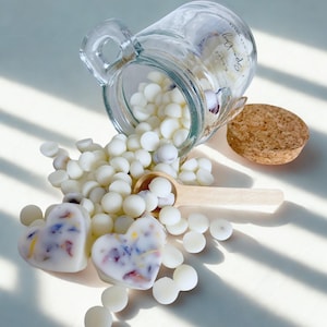 May include: A clear glass jar with a cork lid tipped on its side, spilling small white wax melts onto a surface. Two heart-shaped wax melts with embedded colorful dried flower petals sit in the foreground, accompanied by a small wooden scoop.