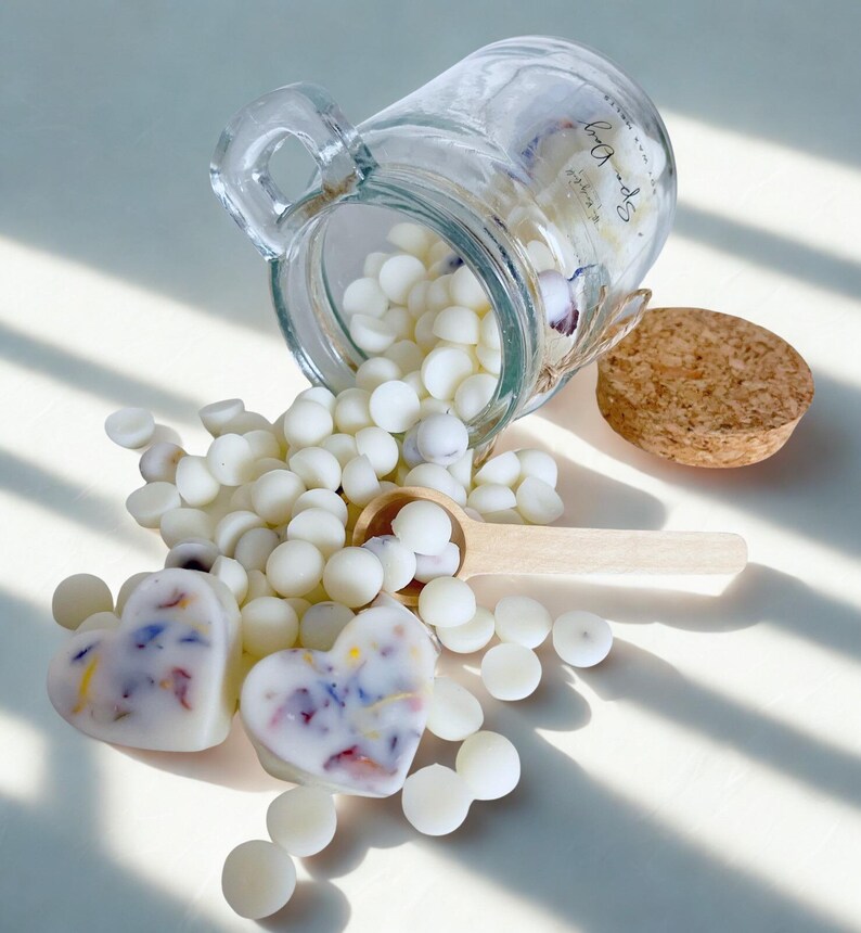 May include: A clear glass jar with a cork lid tipped on its side, spilling small white wax melts onto a surface. Two heart-shaped wax melts with embedded colorful dried flower petals sit in the foreground, accompanied by a small wooden scoop.
