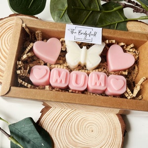 May include: A gift box filled with pink and white wax melts shaped like hearts and the word "MUM". The box is decorated with brown paper and shredded paper.