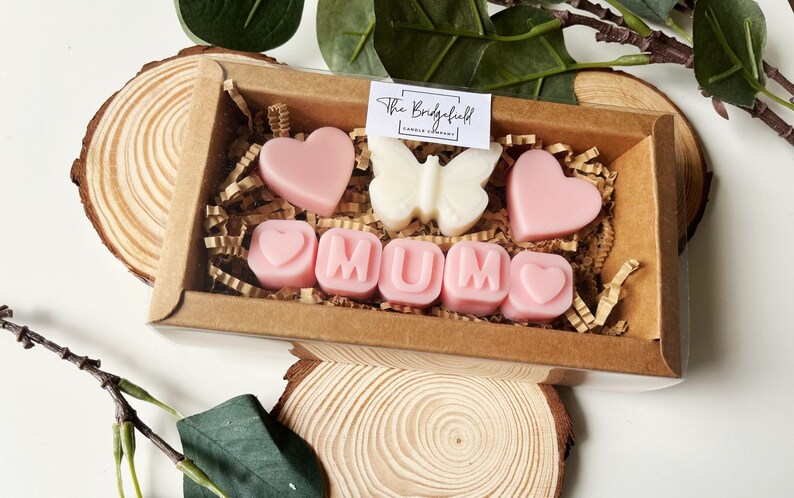 May include: A gift box filled with pink and white wax melts shaped like hearts and the word "MUM". The box is decorated with brown paper and shredded paper.