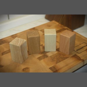 Puede incluir: Cuatro saleros y pimenteros de madera en diferentes tonos de marrón, dispuestos sobre una tabla de cortar de madera.